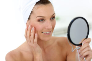 45-year-old woman in bathroom looks at handheld mirror to assess signs of aging on her face